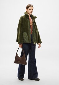 Roheline tekstuuriga liistudega oversize jakk, kantud mustrilise pluusi peal ja kombineeritud tumedate teksapõllega lehvikpükstega ning pruuni käekotiga.