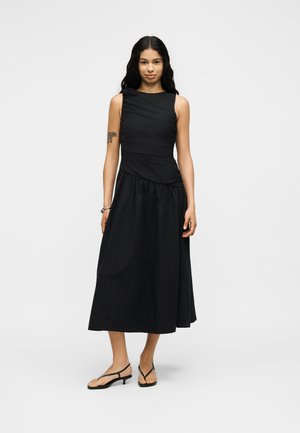 Νεαρή γυναίκα με μακριά μαύρα μαλλιά που φοράει αμάνικο μαύρο midi φόρεμα και μαύρα σανδάλια με χαμηλό τακούνι, στέκεται μπροστά σε ένα απλό φόντο.