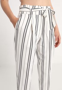 Bílé kalhoty s černými vertikálními pruhy, mají vysoký pas, pásek s mašlí a boční kapsy. Měkká tkanina s hladkým povrchem.