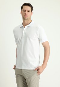 Polo shirt bianco a maniche corte con colletto, patta con due bottoni e texture liscia. Indossato con pantaloni di colore chiaro.