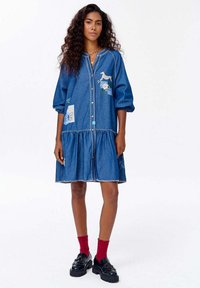 Denimové šaty s gombíkovým zapínaním vpredu, balónovými rukávmi a tiered lemom. Obsahujú vyšívanie, vrátane koňa a kvetinových vzorov, v svetlomodrej farbe.