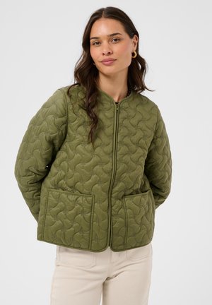 Femme aux longs cheveux bruns portant une veste matelassée vert olive avec des poches plaquées et un pantalon crème, debout devant un fond uni et clair.