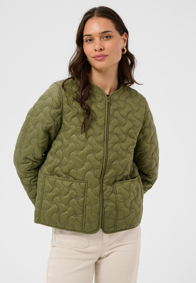 Femme aux longs cheveux bruns portant une veste matelassée vert olive avec des poches plaquées et un pantalon crème, debout devant un fond uni et clair.