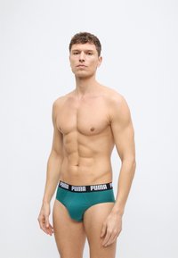 Μυώδης άνδρας όρθιος, φοράει βερμούδες βεραμάν με μαύρη μέση που φέρει την επιγραφή «PUMA», σε απλή ανοιχτόγρια φόντο.