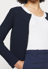 Granatowa, prążkowana kurtka z zamkiem błyskawicznym, okrągłym dekoltem i dopasowanym krojem, w połączeniu z białą, prążkowaną bluzką. Posiada długie rękawy i subtelną teksturę.