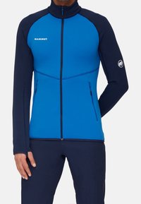 Blauwe fleecejas met marineblauwe accenten, hoge kraag, ritszakken en logo op de borst. Aansluitend ontwerp geschikt voor buitenactiviteiten.