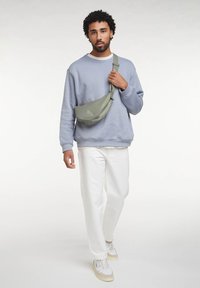 Mann, der einen hellblauen Pullover, weiße Hosen, weiße Turnschuhe und eine grüne Umhängetasche trägt.