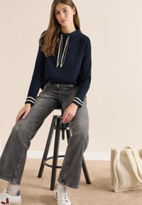 Marineblauer Pullover mit langen Ärmeln und gerippten Ärmeln, Kordelzügen, graue weite Jeans, weiße Sneakers mit roten Akzenten und eine cremefarbene Tote-Bag mit Struktur.