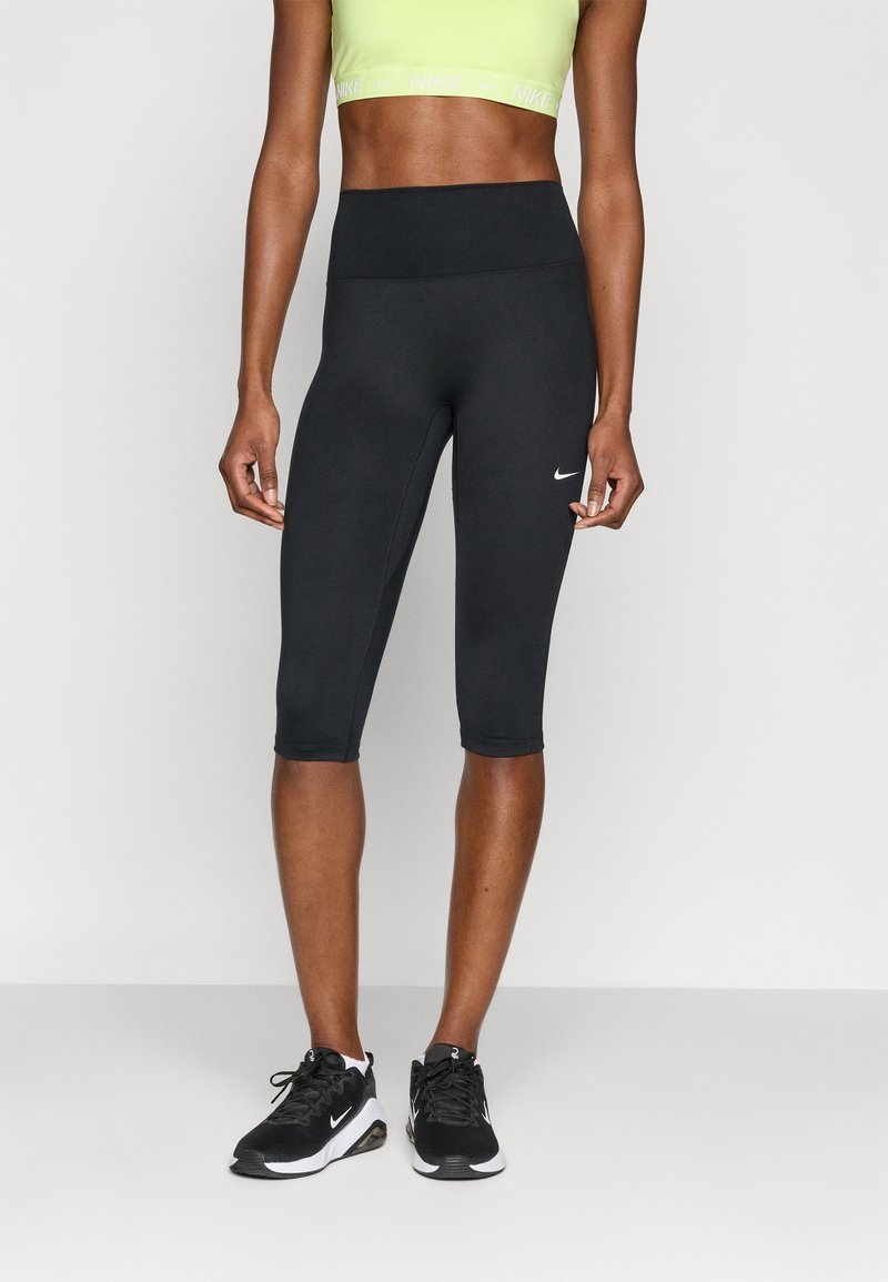 Personne en forme portant des leggings Nike noirs aux genoux, un crop-top de sport vert fluo et des baskets Nike noires, debout sur un fond clair uni.