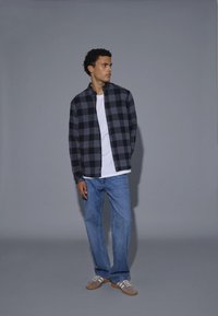 Camicia a quadri nera e grigia sopra una t-shirt bianca, abbinata a jeans azzurri chiari e sneakers grigie. Il tessuto appare morbido e strutturato.