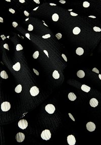 Svart tyg med vita prickar; har en strukturerad yta och ett volangdesign som betonar cirkulära mönster över materialet.