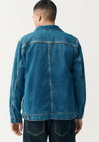 Chaqueta de denim en azul medio con un diseño estructurado, puños abotonados y costuras visibles. Presenta un cuello y un yugo en la parte posterior.