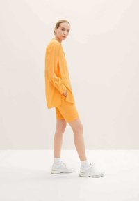 Pomarańczowa, oversize'owa bluzka z marszczonymi rękawami, zestawiona z dopasowanymi szortami. Model nosi białe sportowe buty, na jasnym tle.