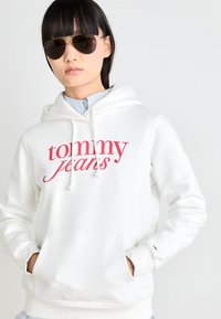 Vit huvtröja med röd "tommy jeans"-logga, frontfickor i känguru-stil, dragsko huva, mjuk textur, bärs över en ljusblå skjorta med krage.