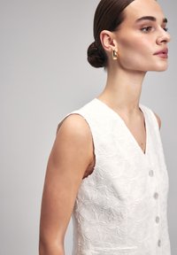 Witte, mouwloze jurk met bloemenborduurwerk, v-hals en knoopdetail aan de voorkant. Model draagt grote gouden oorbellen en heeft een strak naar achteren gekapt kapsel.