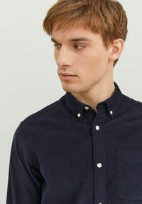 Camisa de pana azul marino con cuello botón, bolsillo frontal y botones blancos. La tela tiene una apariencia texturizada y acanalada.