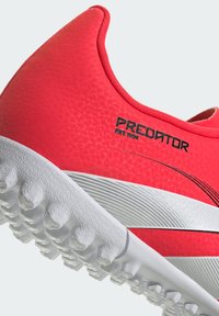 Scarpa da calcio rossa con texture, dal design a taglio basso, con accenti bianchi e tacchetti per terreno solido. Visibile il marchio "PREDATOR EST. 1994".