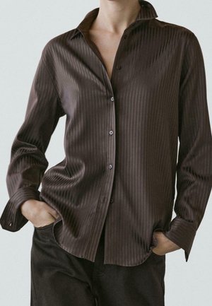 Osoba ubrana w ciemnobrązową, satynową koszulę z długim rękawem i guzikami, z delikatnymi pionowymi paskami, z rękami swobodnie wsuniętymi w kieszenie czarnych spodni.