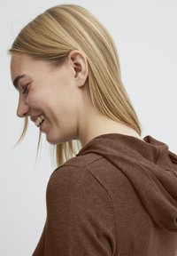 Hoodie marron avec une texture douce, doté d'une capuche et de détails de couture visibles. Modèle présenté de côté avec des cheveux longs.