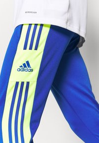 Närbild av en person som bär blå Adidas träningbyxor med neongula ränder och Adidas-logotyp, ihop med en vit Aeroready-tshirt.