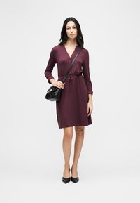 Femme portant une robe bordeaux à manches longues, longueur genou, des escarpins noirs à bouts pointus, et un sac bandoulière noir sur fond blanc.