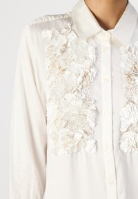 Weiße Bluse mit Kragen, die mit floralen Applikationen und Paillettenverzierungen auf der Vorderseite versehen ist. Glatte Textur und Knopfverschluss.