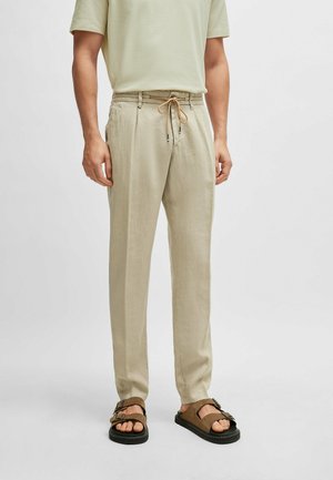 Pantalons en lin beige avec une taille élastique et un cordon de serrage. Coupe droite, tissu texturé et poches latérales. Portés avec des sandales marron.