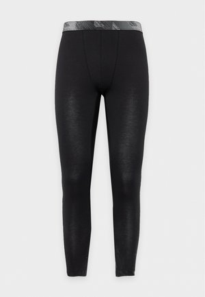 Un homme torse nu porte un legging de compression noir, long jusqu’aux chevilles et doté d’une ceinture à motifs, sur un fond blanc uni.