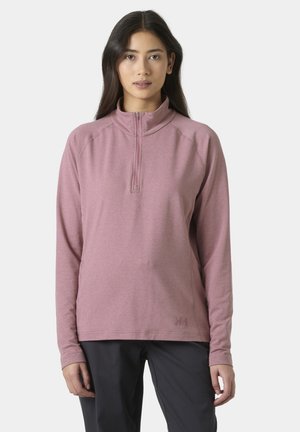 Mujer joven que lleva un jersey malva de manga larga con cremallera hasta la mitad y pantalones negros, parada frente a un fondo claro y sencillo.