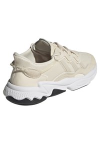 Beige athletic sneaker with mesh and suede upper, textured details, white midsole, and black rubber outsole, featuring a pull tab at the heel.