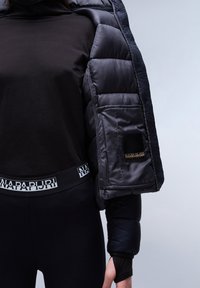 Zwarte langemouwtop gecombineerd met strakke zwarte leggings met een witte logoband. De buitenjas is een donkere gewatteerde jas met textuuraccenten.