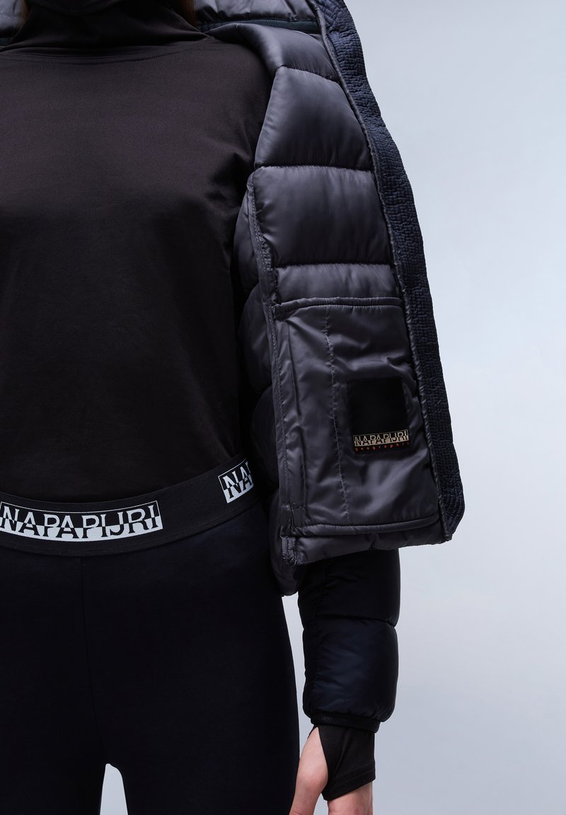 Top nero a maniche lunghe abbinato a leggings neri aderenti con una fascia logo bianca. La giacca esterna è un piumino scuro con accenti texture.