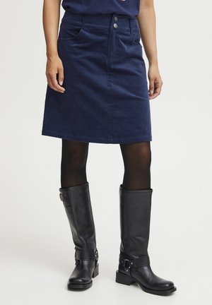 Jupe en velours côtelé bleue marine en forme d'A avec poches avant, fermeture à deux boutons, associée à des bottes en cuir noires montantes.