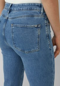 Donkerblauwe denim jeans hebben een hoge tailleband, een achterzak met een studaccent en een slim fit ontwerp. De stof heeft een gladde textuur.