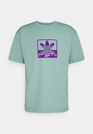 T-shirt vert clair à manches courtes avec un motif violet centré représentant une feuille stylisée et des formes abstraites à l'intérieur d'un cadre carré.