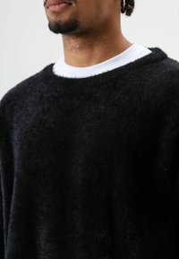 Zwarte, zachte trui met een ronde halslijn, gedragen over een wit shirt met ronde hals. Zachte textuur, ontspannen pasvorm en minimale naden.