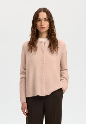Femme aux longs cheveux bouclés portant un cardigan rose pâle boutonné et un pantalon sombre, debout devant un fond clair uni.