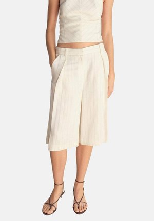 Nainen, jolla on beigejä pinstriper-kuvioituja leveälahkeisia housuhameita taskuilla ja tummanruskeat remmisandaalit yksivärisellä taustalla.