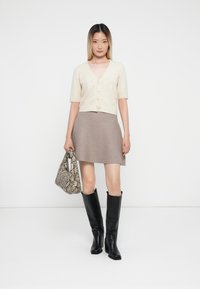 Kremfarget cropped cardigan med korte ermer, parret med et beige flagrende skjørt, svarte knelange støvler og en mønstret slangeskinn-håndveske.