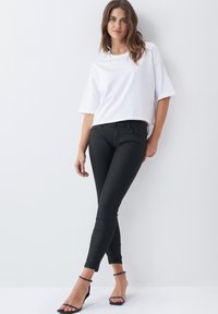 Belo kratki T-shirt z belimi rokavi, v kombinaciji s črnimi oprijetimi jeans hlačami. Model nosi črne sandale z naramnico. Nevtralen ozadje.