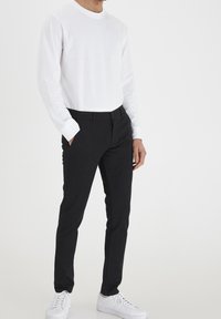 Vit långärmad skjorta, svarta tajta byxor och vita sneakers. Skjortan har rundad hals. Byxorna har sidofickor och en slät tygfinish.