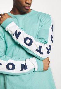 Tričko s dlouhým rukávem v mátově zelené bavlně, s bílými rukávy a textem v námořnické modři. Kulatý výstřih a žebrované manžety.