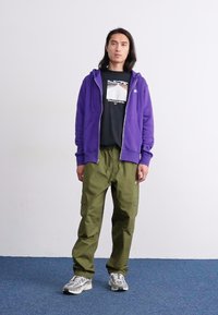 Um hoodie roxo com fecho éclair sobre uma t-shirt preta, combinado com umas calças cargo verde-oliva e ténis cinzentos. Fundo simples com chão azul.