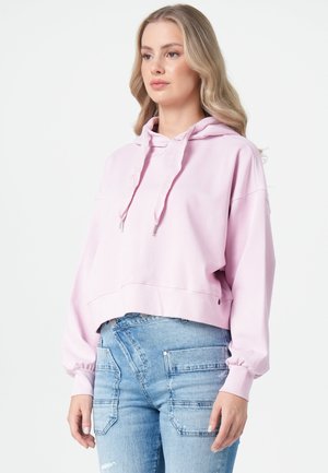 Frau mit welligem blondem Haar, die einen hellrosa Hoodie und blaue Jeans mit großen Vorderaschen trägt, steht vor einem weißen Hintergrund.