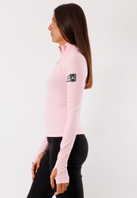 Maglietta a maniche lunghe rosa chiaro con colletto a zip, texture a coste e una toppa nera con logo sulla manica superiore. Stile aderente con fori per i pollici.