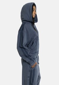 Femeie îmbrăcată într-un hanorac cu glugă albastru închis și pantaloni sport asortați cu dungi laterale, stând în profil cu o mână în buzunar.