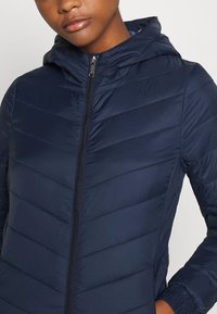 Granatowa kurtka z kapturem, pikowana, z pionowymi szwami, zamkiem błyskawicznym z przodu i elastycznymi mankietami. Lekka, o gładkim, matowym wykończeniu.