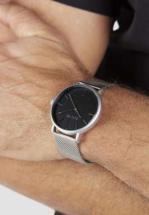 Runde Uhr mit schwarzem Zifferblatt, dünnem silbernem Mesh-Armband, silbernem Metallgehäuse und einfachen Stundenmarkierungen. Getragen an einem Handgelenk mit hellbraunem Haar.