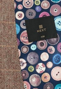 Tissu à chevrons marron à côté d’un tissu coloré à motif de boutons avec une étiquette noire portant la mention "NEXT 40R" cousue dessus.