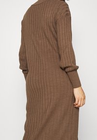Donna che indossa un lungo abito maglione marrone a coste con maniche lunghe, vista di spalle su uno sfondo bianco semplice.